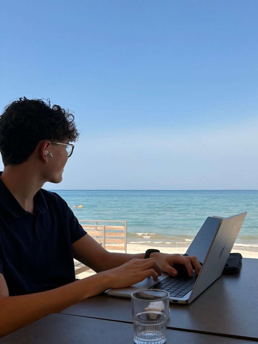 Simon arbeitet am Laptop an einem Tisch mit Blick auf das Meer